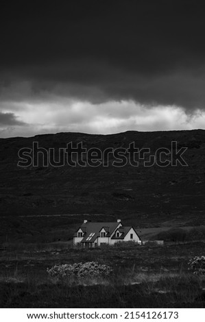 A white house isolated on its own in a field with hills and mountains surrounding it. Stormy weather with dark cloudy sky. Black and white. Concept for loneliness and sadness. Copy space to add text.
