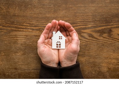 White House Shape In Old Woman Palms On Dark Brown Wooden Table Background. Closeup. Point Of View Shot.