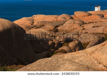 A white house stands tall on a rocky outcrop near the ocean, guiding ships with its beacon. The rugged rocks contrast with the vast expanse of the sea, creating a striking coastal scene.