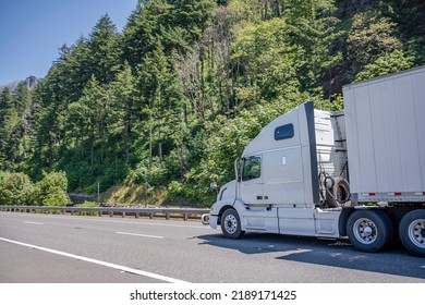 White Industrial Big Rig Semi Truck With Truck Driver Cab Sleep Compartment Transporting Cargo In Dry Van Semi Trailer Running On Summer Highway Road With Forest On Mountain In Columbia Gorge Area