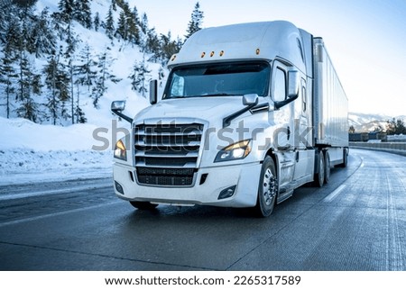 White industrial bonnet big rig semi truck with turned on headlights transporting cargo in dry van semi trailer running on the winter mountain road with lot of snow and ice on the slippery surface