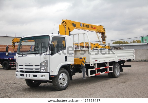 A White Isuzu flatbed truck with yellow crane arm is\
in the parking lot against a gray cloudy sky - Russia, Moscow, 30\
September 2016