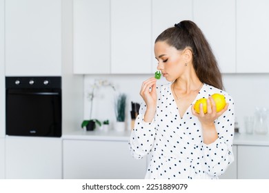 In the white kitchen, a beautiful girl in a white dress with polka dots holds mint in one hand and green lemons in the other. The concept of a cook, nutritionist - Powered by Shutterstock