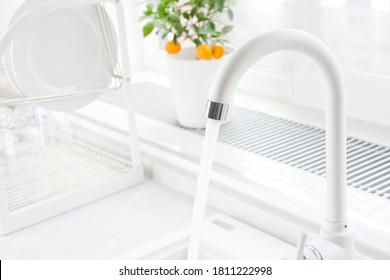 White Kitchen Faucet Running With Blurred Windowsill And Dish Dryer