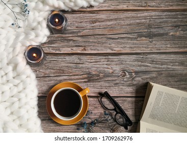 White Knitted Blanket, Coffee Cup, Candles, Reading Glasses And Open Book On Wooden Surface. Top View, From Above, Flat Lay. Still Life, Coziness, Relaxation Concept.