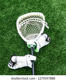 A White Lacrosse Goalie Stick Is Lying On A Green Turf Field With White Gloves Attached.