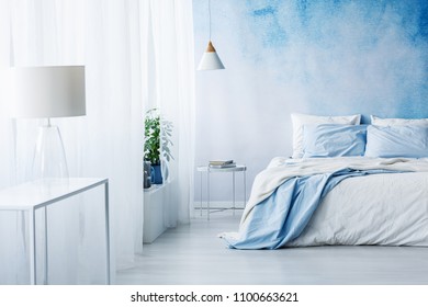 White Lamp On A Table In Bright Blue Bedroom Interior With Bed Against Ombre Wall