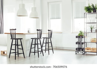 White Lamps Above Kitchen Island Under A Window With Black Bar Stools In Bright Room With Plants