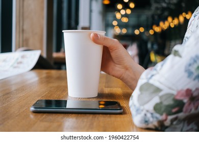 White Large Paper Cup Of Tea On A Table In A Cafe. A Woman's Hand Holds The Cup, A Smartphone Lies Next To It. The Concept Of A Break, Rest In A Cafe. Copyspace.