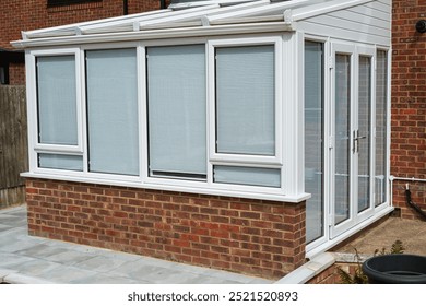 White lean to conservatory with blinds on all windows and doors, seen from the outside on a sunny day. 