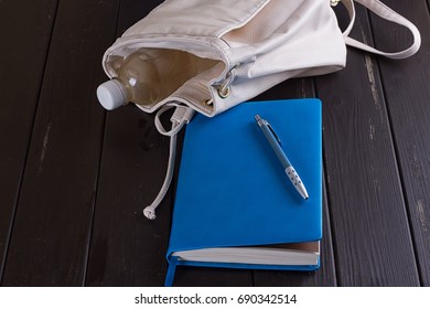 White leather backpack, blue notebook and pen on a black wooden background