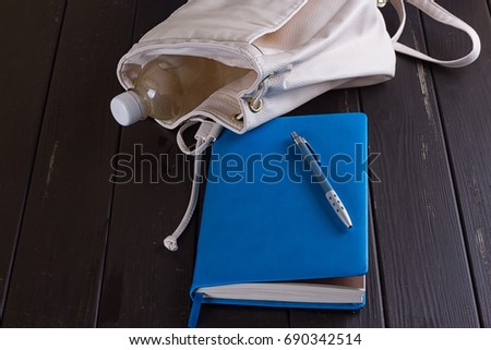 White leather backpack, blue notebook and pen on a black wooden background
