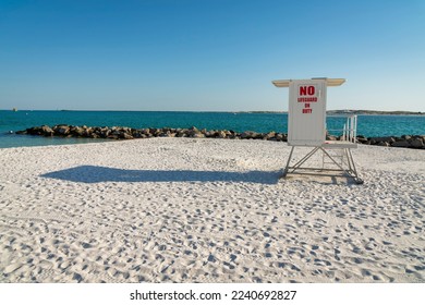 White lifeguard tower with painted red No Lifeguard On Duty lettering sign on the door- Destin, FL. Lifeguard tower on a white sand seashore with rocks near the blue ocean water against the sky.