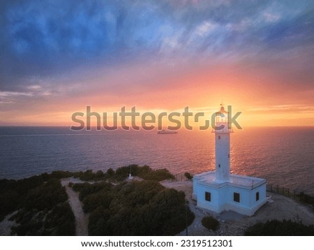 White lighthouse on Cape Ducato with sun just in glassed-in housing at the top of a lighthouse tower, sunset and cruise ship in background, very colorful and epic moment. Lefkada Island, Greece.