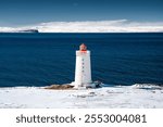 White lighthouse with red top stands on a snowy coastline with deep blue water in the background under a clear sky.