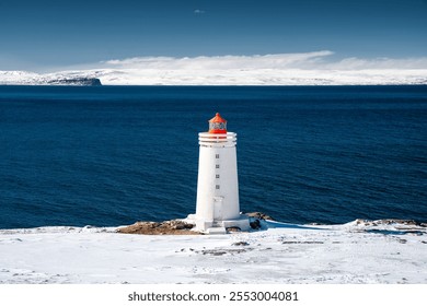 White lighthouse with red top stands on a snowy coastline with deep blue water in the background under a clear sky. - Powered by Shutterstock