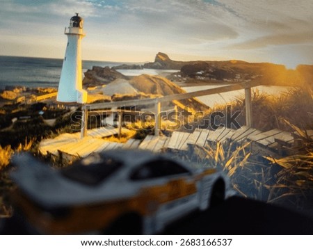 A white lighthouse rises gracefully by the seaside at sunset, with a winding wooden path and the silhouette of a miniature car in the foreground adding an artistic touch.