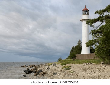 A white lighthouse, rocky shoreline, surrounded by lush greenery. The cloudy sky looms overhead, adding a sense of drama to the serene coastal scene.  Estonian island. Sea, rocky beach, summer evening - Powered by Shutterstock