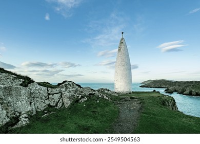 A white lighthouse stands out on the rocky coastline, surrounded by grass and cliffs. The calm ocean stretches out under a clear blue sky, creating a sense of tranquillity. - Powered by Shutterstock