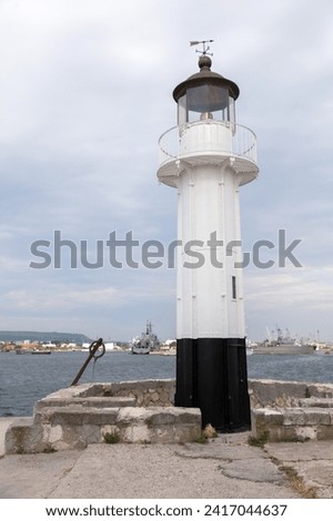 White lighthouse tower with black base is under blue sky on a daytime, vertical photo. Port of Varna, Bulgaria