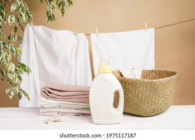 White linen is dried on a clothesline against the background of a beige wall with a houseplant. A stack of colored laundry, a laundry basket, bottles of detergent and fabric softener are on the table. - Shutterstock ID 1975117187