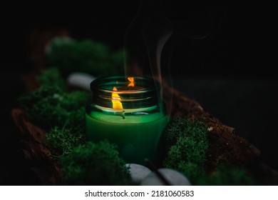 A white lit candle in a glass jar with flowing smoke, green moss and gray stones in the bark of a tree lies diagonally against a black background, close-up side view. Esoteric concept, dark style. - Powered by Shutterstock