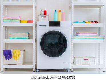 White Luandry Machine With Washing Powder
 Liquid And Equipments On Side Shelf With Clean Colorful Clothes.