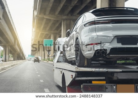 A white luxury car being transported on a tow truck under an elevated highway on a sunny day.
