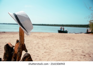 White Male Hat On Old Tree Log At Sunny Sand Beach With Blurred Fishermen In The Boat On Background. Copy Space