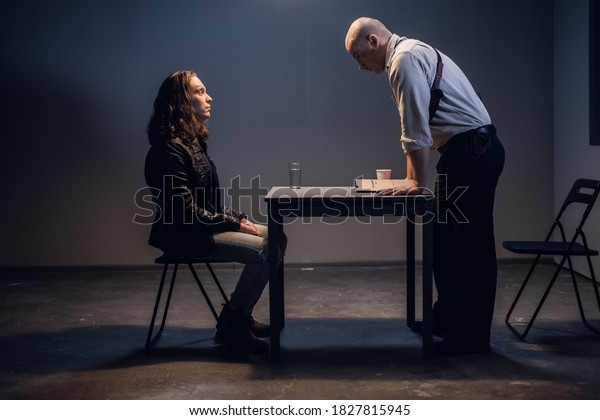 A white male police detective talks to a\
suspect in a drug trafficking case who is sitting in an\
interrogation room in\
handcuffs