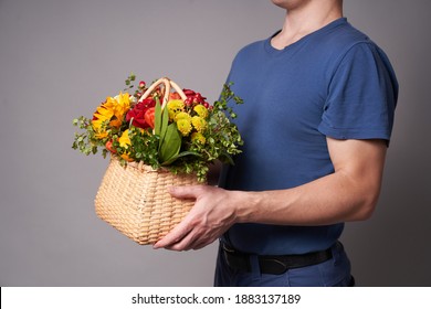 A White Man In A Blue T-shirt Holds Out A Basket Of Flowers With A Copy Space On A Grey Background, Flower Delivery Or A Gift For Holiday