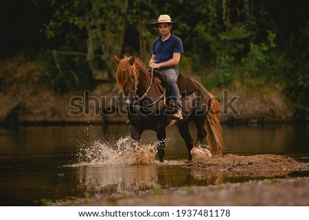 White man riding a horse inside a river, wearing a straw hat, with his horse in a saddle and throwing water into the air.
