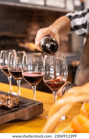 A white man serving the last of four glasses of red wine in a row next to a gaucho traditional barbecue on a rustic wooden board in a home or restaurant to have a nice lunch or dinner with some guests