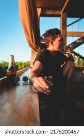 White Man Sitting At Rooftop Bar Holding A Tequila Shot With Wood Furniture And Beer Bottle And Blue Sky In The Background In Tulum, Quintana Roo, Mexico On September 12th 2021