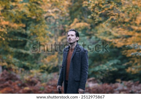 A white man stands in Epping Forest, London, looking upward with a contemplative expression amidst a backdrop of vibrant autumn foliage