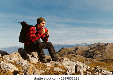 white man with tattoos, red clothes and backpack having a drink (coffee, water or tea) on a mountain peak while contemplating the mountain scenery. hiker taking a break. travel and adventure. - Powered by Shutterstock