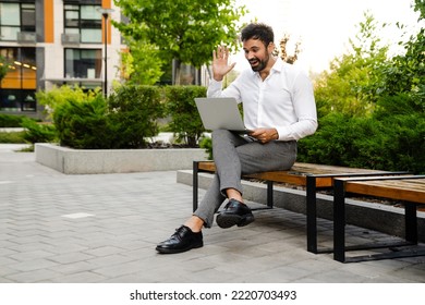 White Man Waving Hand And Using Laptop While Sitting On Bench Outdoors