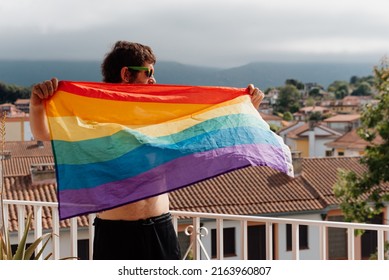 White Man Waving The Rainbow Flag Of Lgbt Pride On The Terrace Of His House. Real People At Home.