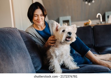 White Mature Woman Petting Her Dog While Sitting On Couch At Home