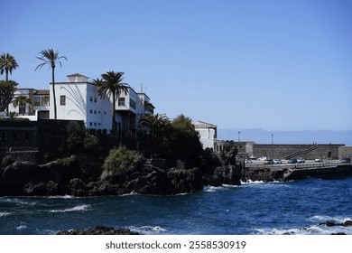 White Mediterranean-style buildings with palm trees and terraces stand on a rugged rocky coastline, surrounded by the turquoise waves of the ocean, with a clear blue sky stretching to the horizon and  - Powered by Shutterstock