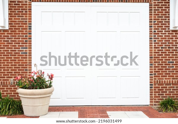 A white metal garage panel door in a red brick house.\
There\'s a large clay pot of flowers in from of the door. The\
entrance to the building has bricks on the ground with two rows of\
white blocks.  