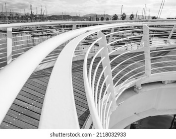 A white metal handrail curves in an arc against the backdrop of a marina with yachts and palm trees. Black and white photo