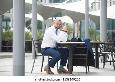 White Middle Age Businessman Wearing A Tie With Glasses & Beard Sitting At An Outdoor Cafe While On The Phone With A Serious Or Stressed Look On His Face  