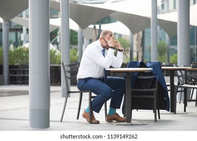 White Middle Age Businessman Wearing A Tie With Glasses & Beard Sitting At An Outdoor Cafe While On The Phone With A Serious Or Stressed Look On His Face  