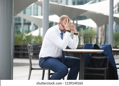 White Middle Age Businessman Wearing A Tie With Glasses & Beard Sitting At An Outdoor Cafe While On The Phone With A Serious Or Stressed Look On His Face  