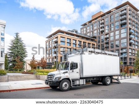 White middle class relocation rig day cab semi truck tractor with long spacious box trailer standing on the urban city street with multilevel apartment and office buildings unloading delivered goods