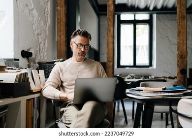 White middle-aged man in earphones using laptop computer while sitting in modern office - Shutterstock ID 2237049729