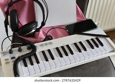 White MIDI keyboard controller, black headphones, smartphone, notebook, pen on pink fabric surface close-up