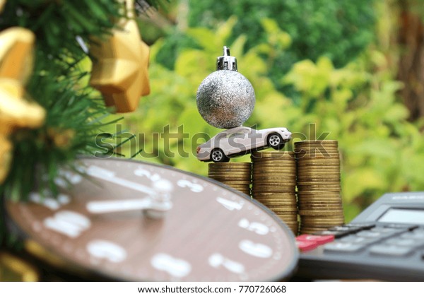 White miniature car carry glitter ball drives on roll\
ladder f gold coin money, calculator and blur wood clock placed\
near Christmas tree decorated with gold star in blurred natural\
garden  