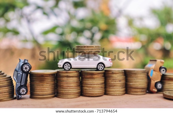 White miniature car carry gold coins on rolls coins and\
toy cars stuck between pile of money on wood table in blur natural\
tree 